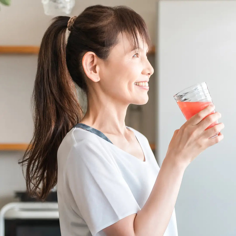 Profile of a smiling middle-aged Asian woman holding a glass of pink liquid (juice or smoothie) in a bright kitchen.