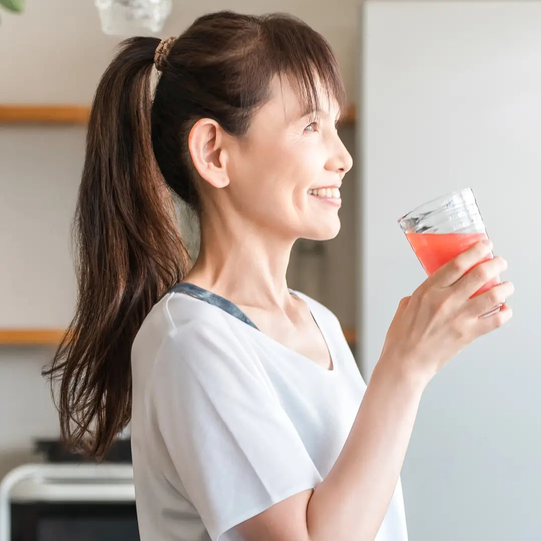 Profile of a smiling middle-aged Asian woman holding a glass of pink liquid (juice or smoothie) in a bright kitchen.