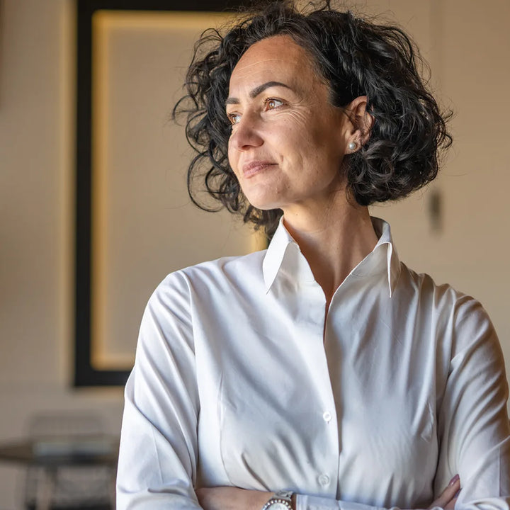 Woman with dark curly hair wearing a white shirt, looking to the side with a confident expression in a naturally lit office setting.